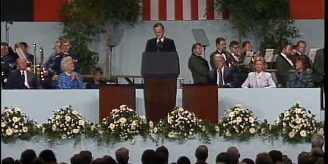 George H.W. Bush delivering speech at a lectern to a crowd with flowers, an American flag, teleprompters, and people sitting behind him
